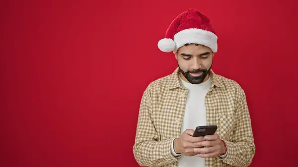 Junger Hispanischer Mann Trägt Weihnachtsmütze Mit Smartphone Vor Isoliertem Roten — Stockfoto