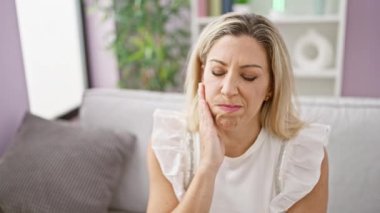 Kanepede oturan genç sarışın kadın evinde diş ağrısı çekiyor.