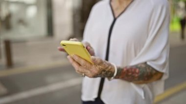 Ciddi bir ifadeyle kıdemli, kır saçlı, güneş gözlüklü, şehir güneşinin altında duruyor, cep telefonuna dalmış, sokakta internet mesajı yazıyor..