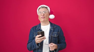 Noel Baba şapkası takan bir adamın portresi akıllı telefon ve kredi kartı kullanarak kırmızı arka planda poz veriyor.