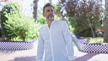 Endişeli, gri saçlı genç İspanyol adam şehir parkındaki kol saatinin zamanını endişeyle kontrol ediyor. Yakışıklı bir yüz, geç kalma ihtimalinin ciddi kaygısını ortaya çıkarır..