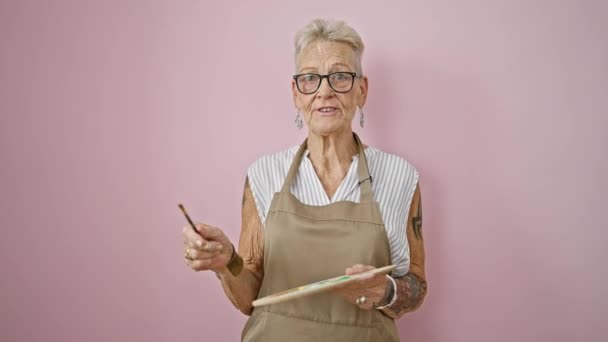 自信を持って灰色の髪のシニア女性アーティスト 喜んで話す 孤立したピンクの背景図面の上にペンキブラシとパレットと立って — ストック動画