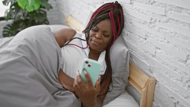 アフリカ系アメリカ人女性が寝室の快適さでスマートフォンを使用し 落ち着かない朝の雰囲気でベッドに座っている — ストック動画