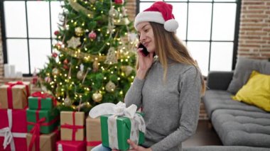 Genç ve güzel bir kadın telefonda konuşuyor ve oturma odasında Noel hediyesini tutuyor..