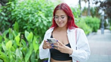 Neşeli genç kızıl saçlı kadın akıllı telefon kullanıyor, parkın yemyeşil yeşili içinde mesaj atıyor, doğayı ve modern teknolojiyi kucaklıyor.