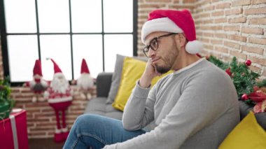 Genç, yakışıklı, sakallı, kırmızı şapkalı adam Noel ağacının önündeki koltukta oturuyor ve sıkılmış görünüyor.