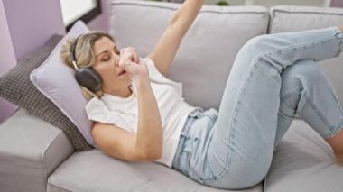 Genç sarışın kadın evde dans ederken kanepede müzik dinliyor.