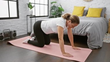 Çekici, genç, sarışın bir kadın yatak odasında uzanırken gülümsüyor ve sabah sporu için ısınıyor.