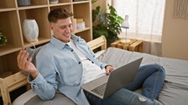 Kafkasyalı genç adam laptopuyla video görüşmesi yapıyor. Yatak odasındaki yatakta oturuyor.