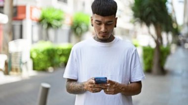 Gülümseyen genç latin adam açık havada akıllı telefonuyla mutlu bir şekilde mesajlaşıyor, yakışıklı dövmeli bir yetişkin şehir caddesinde neşeli bir dijital sohbete dalmış..