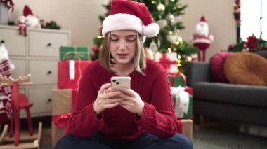 Kırmızı Noel Baba şapkalı genç bir kadın cep telefonu kullanıyor ve Noel 'de evde gülümsüyor.