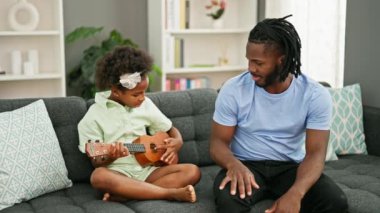 Afrikalı Amerikalı baba ve kız kanepede oturup evde ukulele çalmayı öğretiyor.