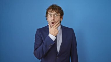 Şaşkınlık içindeki takım elbiseli genç adam şok ve inanmazlığını ifade ediyor, elleri çenesinde, dışlanmış mavi arka planda şaşkın bir ifade.