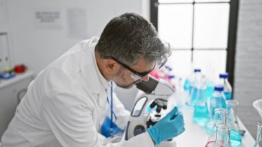 Çekici gri saçlı İspanyol bir adam, kendini adamış bir bilim adamı, laboratuarda tıbbi bir deney üzerinde çalışıyor, güvenlik eldivenleri takılı, mikroskoptan dikkatle bakıyor,.