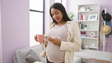 Tasasız genç hamile kadın, sabah kahvesinden zevk alırken gülümsüyor, rahat oturma odasında akıllı telefondan mesajlaşıyor.