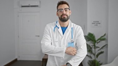 Kendine güvenen genç İspanyol bir adam, klinik bekleme odasında kolları çapraz duran gülümseyen bir doktor, pozitif cazibe ve profesyonel sağlık becerisi yayıyor.