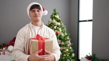 Noel Baba şapkalı bir adam elinde süslü Noel ağacıyla elinde hediyesiyle evde duruyor.