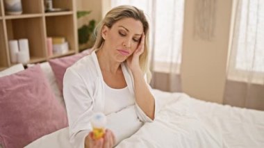 Genç sarışın kadın elinde ilaç şişesiyle baş ağrısı çekiyor. Yatak odasında oturuyor.