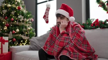 Noel Baba şapkalı genç adam kırmızı ekoseli koltukta oturuyor, sıkılmış ve üzgün görünüyor.