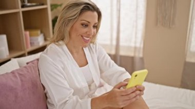 Yatak odasında akıllı telefon kullanan genç sarışın kadın.