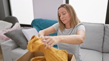 Çekici genç sarışın kadın evinde oturma odasındaki kanepede rahatça otururken bir paketin içine neşe içinde kıyafet dolduruyor.