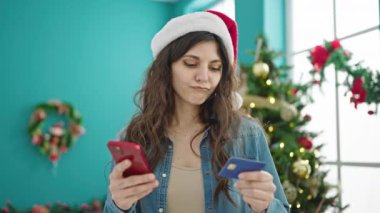 Genç ve güzel İspanyol bir kadın Noel ağacının yanında akıllı telefon ve kredi kartıyla alışveriş yapıyor.