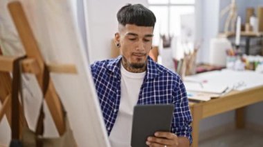 Kendine güvenen genç bir Latin, dövmeli bir sanatçı, dokunmatik defterine çizim yaparken gülümsüyor ve kapalı alanda resim sınıfında oturuyor.