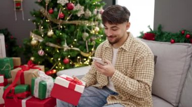 Yakışıklı, mutlu, genç bir adam Noel hediyesini tutuyor ve evde akıllı telefon kullanıyor. 