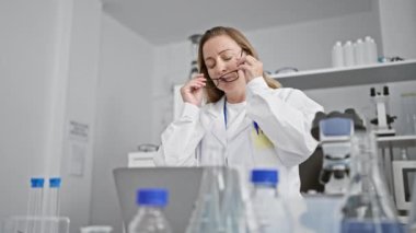 Gülümseyen, kendine güvenen, gözlüklü genç sarışın bilim adamı kusursuz laboratuvar ortamında dizüstü bilgisayar kullanıyor.