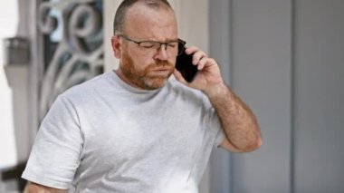 Yakışıklı, orta yaşlı, beyaz bir adam. Güneşli şehir sokağında duruyor. Havalı akıllı telefonuyla ciddi bir telefon konuşması yapıyor.