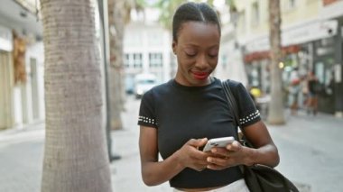 Neşeli Afro-Amerikalı kadın, şehir caddesinde dikilirken telefonuyla mutlu mesut mesajlaşıyor.