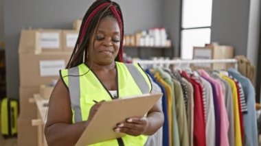 Afro-Amerikan kadın, neşeli bir şekilde gülümsüyor, bir hayır kurumunda gönüllü olarak çalışıyor, yansıtıcı yeleğiyle duruyor, bağış kutularıyla çevrili bir pano kontrol listesine yazılmış.