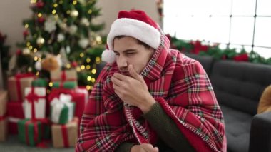 Genç hasta adam battaniyeye sarılıp süslenmiş oturma odasında Noel şapkası takıyor.