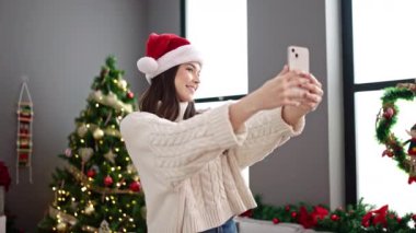 Noel Baba şapkalı mutlu genç bir kadın süslü Noel ağacında cep telefonuyla selfie çekiyor. Noel 'i evde kutluyoruz. xmas kutlaması.