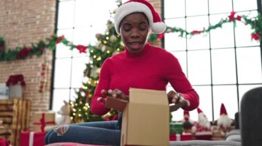 Noel Baba şapkalı mutlu Afrikalı Amerikalı kadın oturma odasında Noel hediyesini açıyor ve heyecanlı görünüyor. 