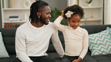 Afro-Amerikan baba ve kızı gülümseyerek koltukta oturmuş evde konuşuyorlar.