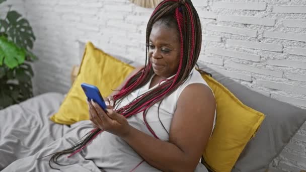 ベッドの上に座っている美しいアフリカ系アメリカ人女性 リラックス オンラインで勝利を祝う 携帯電話で スマートフォンを使用してメッセージを入力 彼女の寝室の屋内でのライフスタイルを楽しむ — ストック動画