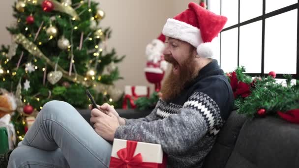 Szczęśliwy Człowiek Kapeluszu Świętego Mikołaja Siedzi Kanapie Rozmawia Smartfonie — Wideo stockowe