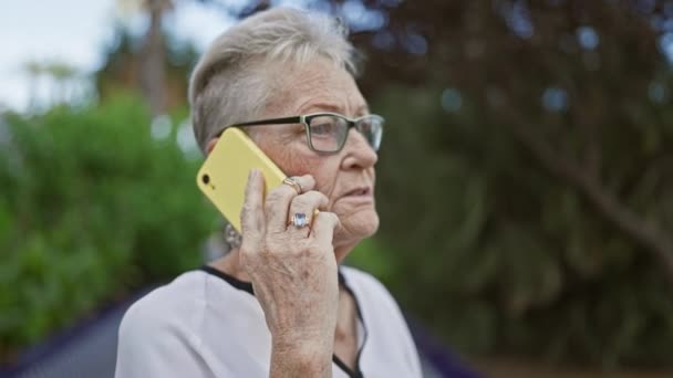 在阳光普照的公园里 一位冷酷 白发苍苍的老妇人在外面认真地和智能手机交谈时 脸上流露出一种随意而时尚的生活方式 神魂颠倒的神情 — 图库视频影像