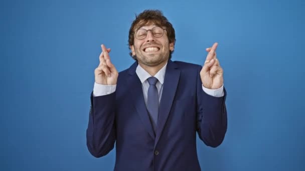 ビジネスススーツの陽気な若者 運と希望のために指を交差し 目を閉じる 単離された青い背景に対する希望的な兆候をジェスチャー — ストック動画