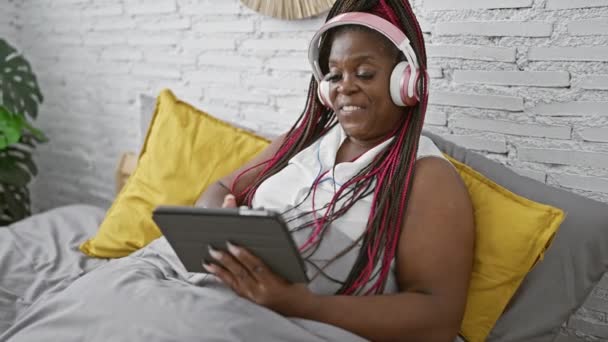 穿着辫子 头戴触摸板装置 头戴耳机 快乐地躺在卧室里的快乐的非洲裔美国女人 沉浸在一个网上世界里 — 图库视频影像