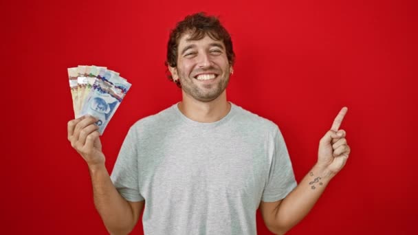 カナダのドル紙幣を喜んで持っている若者が 赤い背景に孤立したフレンドリーな笑顔で側を指摘することを願っています — ストック動画