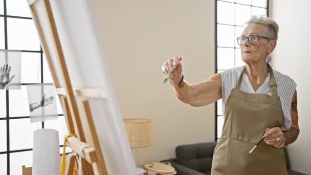 エプロンの真剣で灰色の髪のシニア女性は 真剣に屋内スタジオでアートを描くことに集中し キャンバス上の彼女の成熟した創造性を体現しています — ストック動画