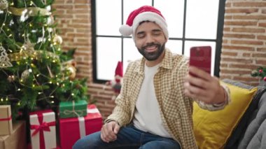 Noel Baba şapkalı genç adam evde yeni yılı kutluyor, Noel ağacı ve arka planda hediyelerle selfie çekiyor. 