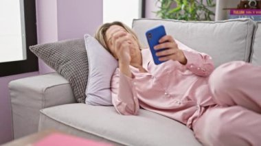 Kanepede uzanmış akıllı telefon kullanan genç sarışın kadın. Evde ciddi bir yüzü var.