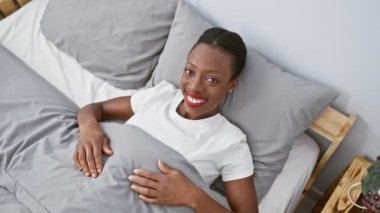 Neşeli Afrikalı Amerikalı kadın yatak odasındaki rahatlatıcı bir yerden seni işaret ediyor. Yatağında uzanırken yüzünde kendine güvenen bir gülümseme beliriyor.!