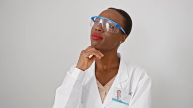 Kafası karışmış Afro-Amerikan kadın bilim adamı üniforması, çenesinde parmak, düşüncelere dalmış, izole edilmiş beyaz arka planda hayret içinde.