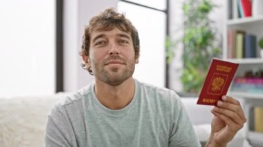 Neşeli genç adam oturma odasını turluyor. Mutlulukla elinde Rus pasaportu, rahat bir şekilde kanepede oturuyor.