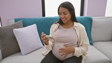 Neşeli genç hamile kadın evinde rahat bir şekilde rahatlıyor, akıllı telefondan video izlerken gülümsüyor ve karnına sevgiyle masaj yapıyor.
