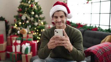Oturma odasındaki Noel ağacında otururken akıllı telefon kullanan mutlu genç adam.